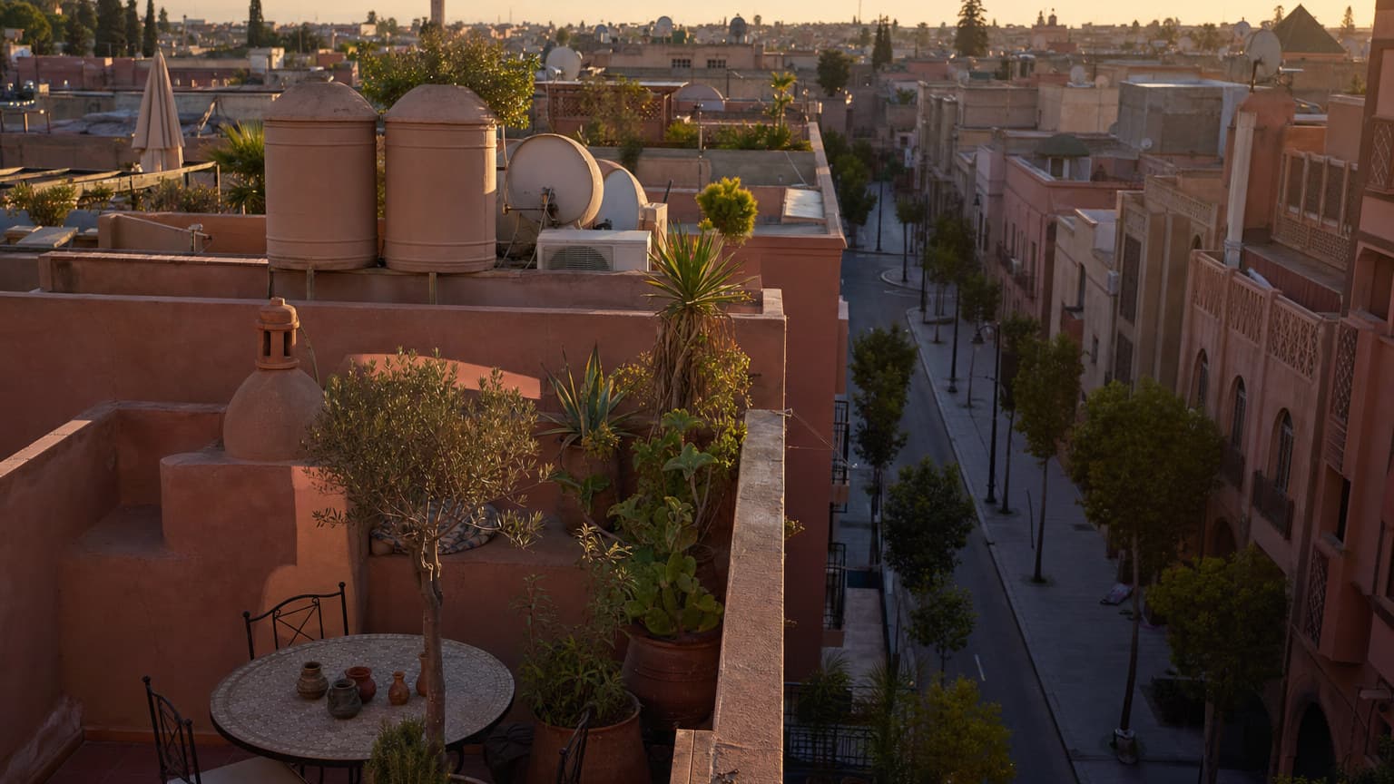 Terracotta rooftop terrace of a Moroccan apartment building with water tanks and satellite dishes in warm late-afternoon light, overlooking a quiet residential street