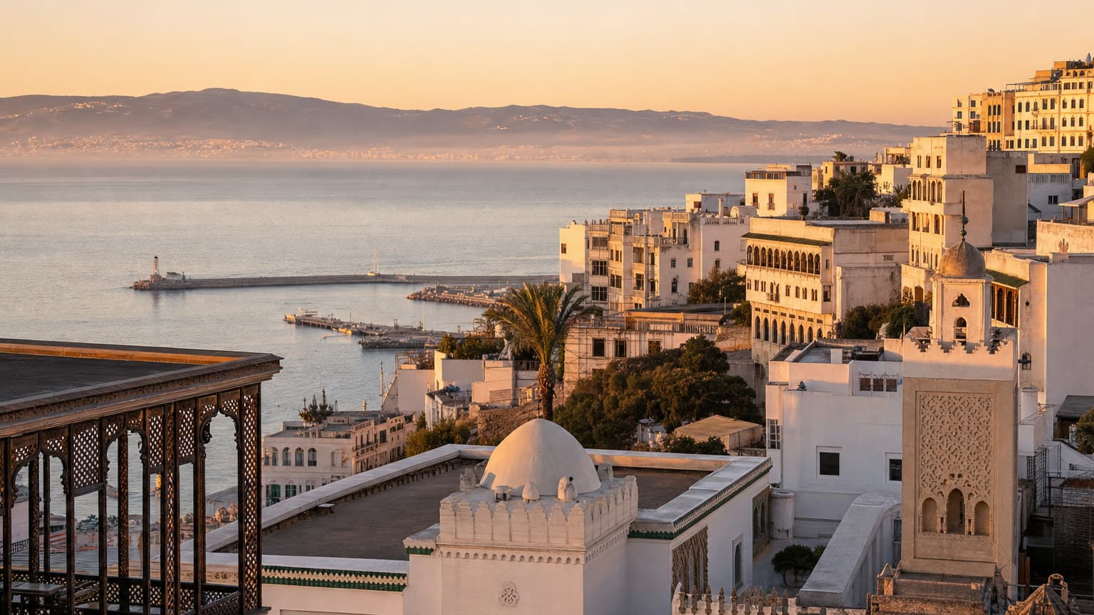 Whitewashed buildings climbing a hillside above the bay of Tangier at golden hour, with the Strait of Gibraltar visible in the distance